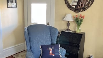 Un fauteuil bleu agrémenté d’un coussin décoratif, un meuble noir, un miroir et un vase de fleurs, le tout posé sur un plancher en bois.