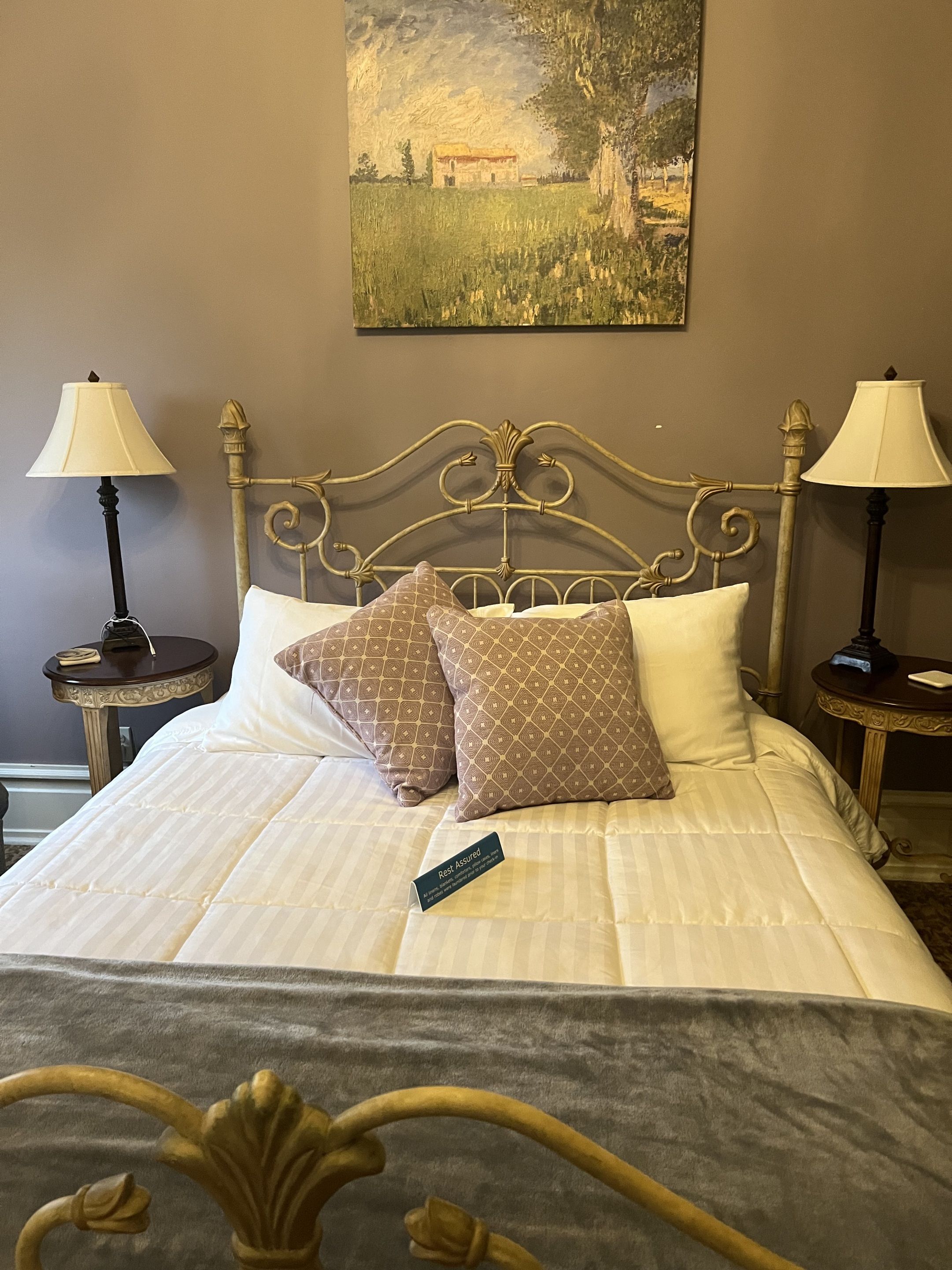 Un lit bien fait, avec une pancarte indiquant qu’il est prêt pour les invités, deux tables de chevet avec des lampes et un tableau accroché au mur.