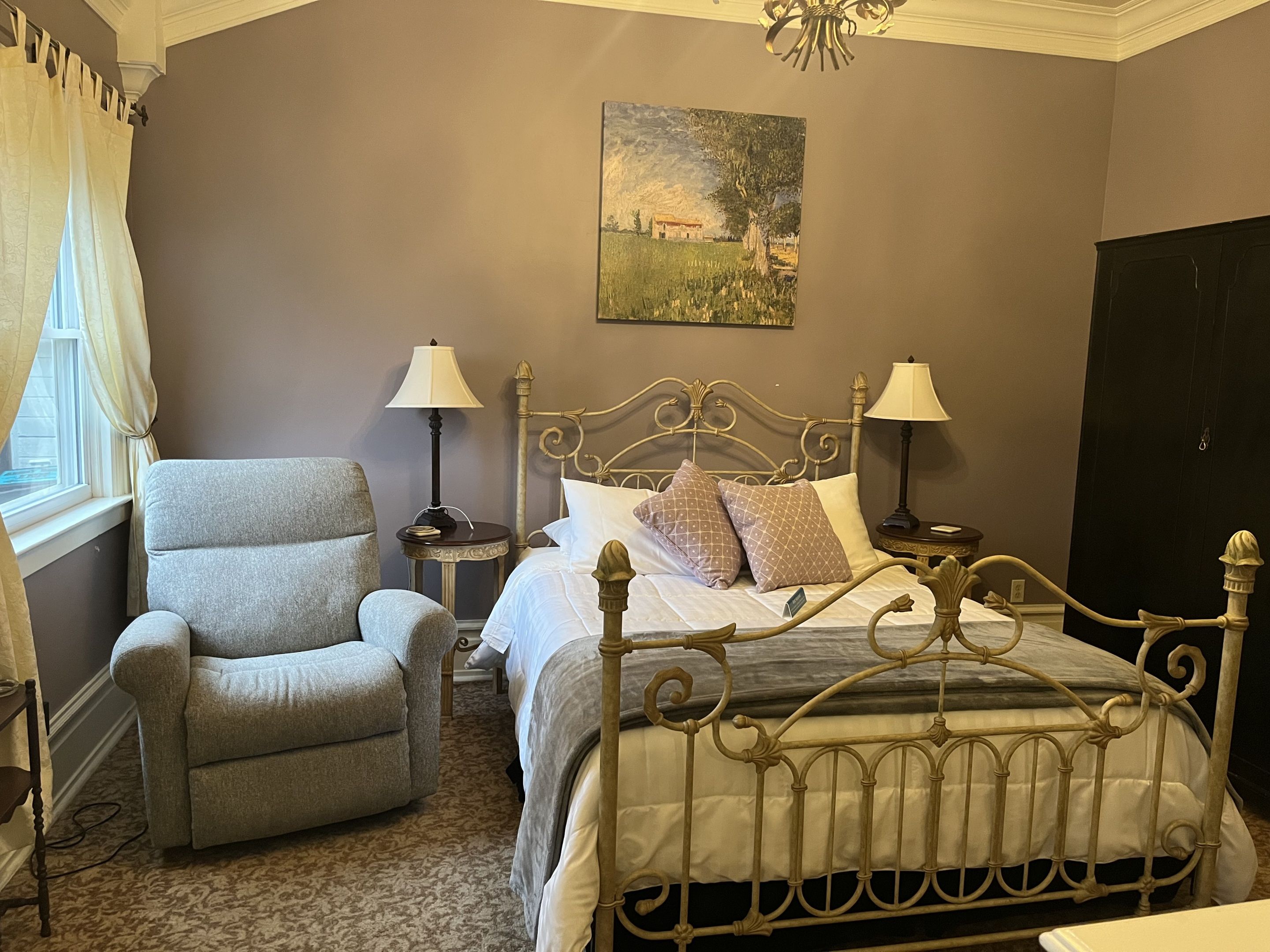 A bedroom with a metal bed frame, a grey armchair, two bedside tables with lamps, a painting on the wall, and a window with curtains.
