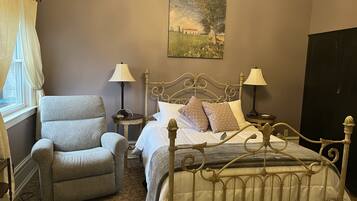 A bedroom with a metal bed frame, a grey armchair, two bedside tables with lamps, a painting on the wall, and a window with curtains.