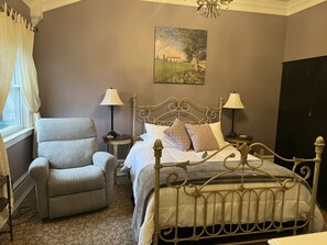A bedroom with a metal bed frame, a grey armchair, two bedside tables with lamps, a painting on the wall, and a window with curtains. - Abbey House Inn (Winters)