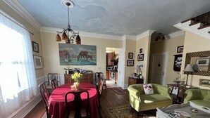 Une salle à manger avec une nappe rouge, des chaises et un lustre. Un tableau orne le mur, et on aperçoit une cuisine au fond.