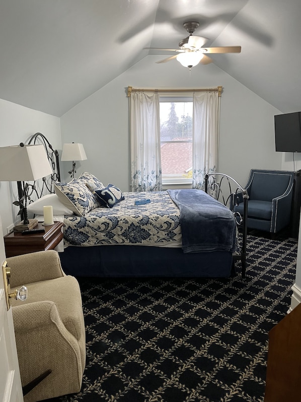A bedroom with a bed, two bedside tables, a chair, a ceiling fan, a window with curtains, and a television on the wall. - Abbey House Inn (Winters)
