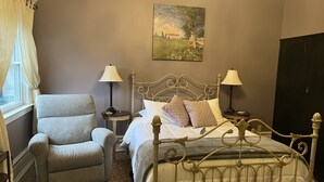 Une chambre à coucher avec un lit en métal, un fauteuil gris, deux tables de chevet avec des lampes, un tableau au mur et une fenêtre avec des rideaux.