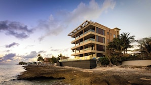 A modern multi-story building with balconies, located on a rocky shoreline with a view of the ocean. - SeaDreams 1: Ocean Condos w/Pool. Jacuzzi & Rooftop Patio; Stay a Week, Save 20% (West Bay)