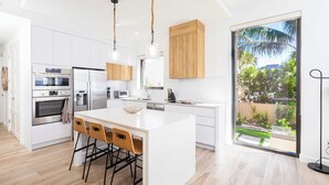 A modern kitchen with white cabinetry, stainless steel appliances, a large island with bar stools, and a sliding glass door to a garden. - SeaDreams 1: Ocean Condos w/Pool. Jacuzzi & Rooftop Patio; Stay a Week, Save 20% (West Bay)