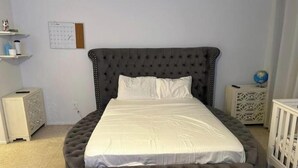Une chambre à coucher avec un grand lit, un pouf rond, une table de chevet, une commode, un globe terrestre et un calendrier accroché au mur.