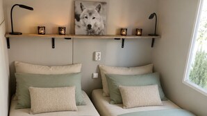 Un dormitorio con dos camas, un estante en la pared con objetos decorativos y una ventana que deja entrar luz natural.