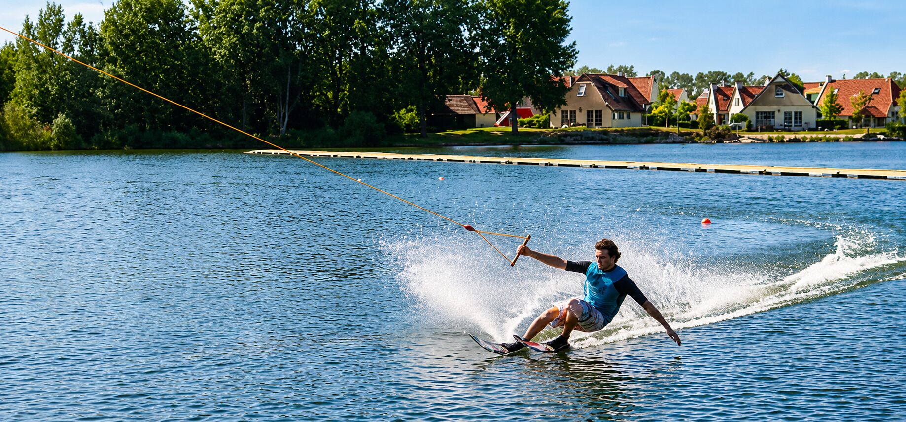 Una persona practica wakeboard en un lago tranquilo con casas al fondo.