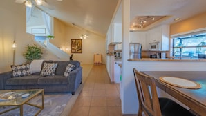 A modern living room with a gray sofa, a glass coffee table, and a dining area with a wooden table and chairs. A kitchen with white cabinets and appliances is visible in the background. - Symphony Suite (Paso Robles)