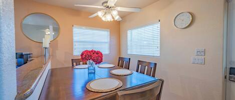 A dining room with a wooden table, chairs, a ceiling fan, and a wall clock.
