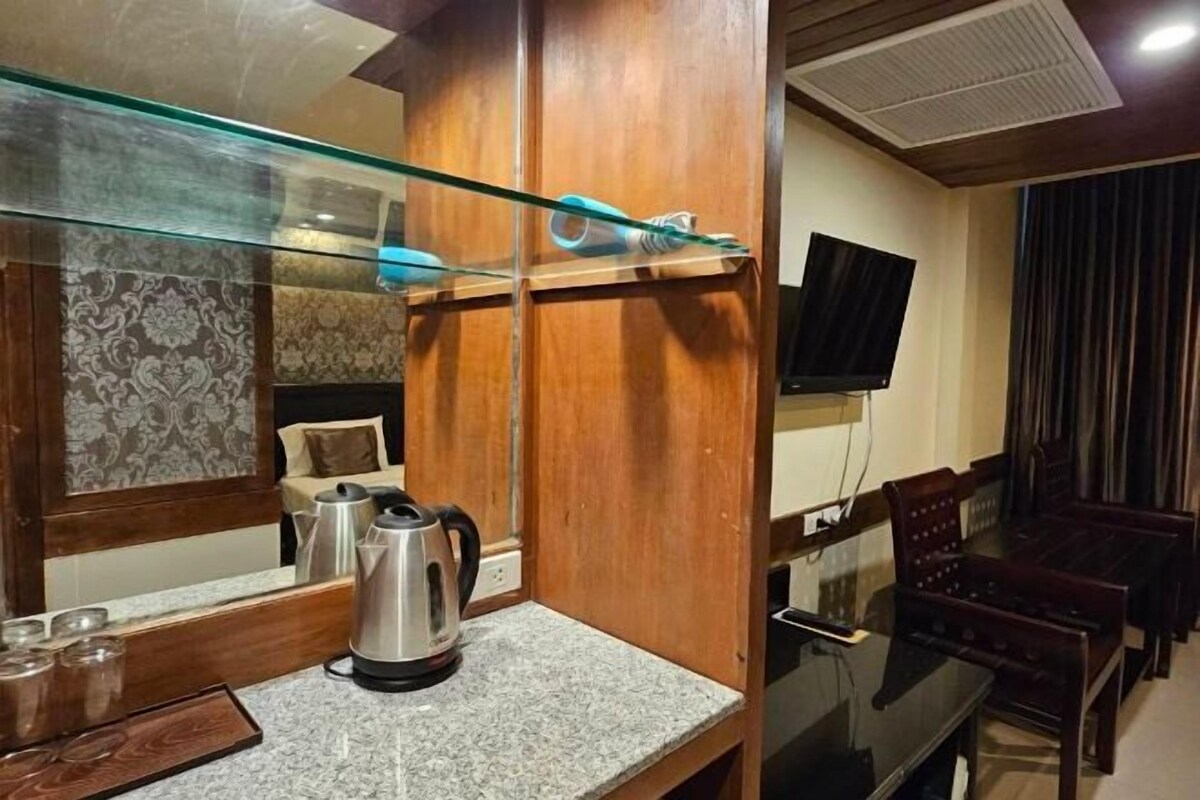 A hotel room with a wooden cabinet, a glass shelf, a kettle, a TV, and a dining area with chairs and a table.