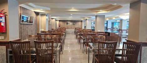 Une salle à manger d’hôtel avec des rangées de tables et de chaises en bois, un extincteur sur le côté gauche et un téléviseur accroché au mur.