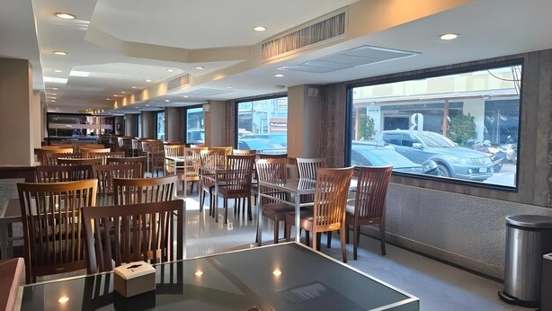 Un espace de restauration moderne avec des chaises et des tables en bois, de grandes fenêtres donnant sur la rue et une poubelle visible.