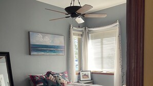 Un dormitorio con cama, ventilador de techo, ventana con persianas y un cuadro en la pared.