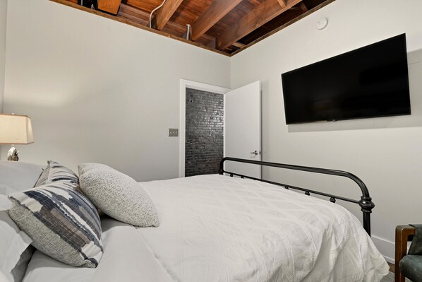 Un dormitorio con cama, almohadas, televisor, pared de ladrillo y puerta.