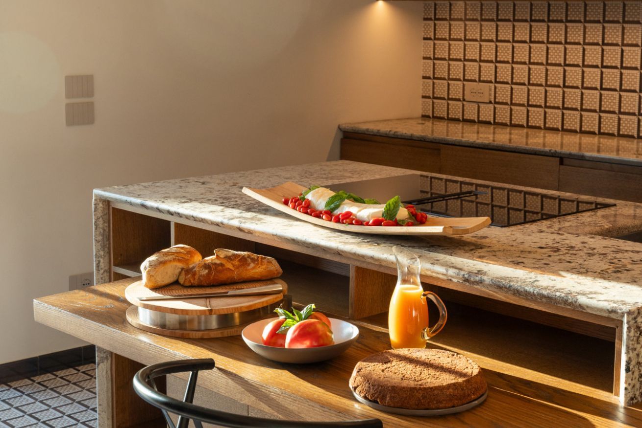 A breakfast setup with bread, tomatoes, a plate of food, and a jug of orange juice on a kitchen counter.