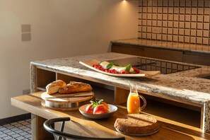 Une table de petit-déjeuner avec du pain, des tomates, une assiette de nourriture et une carafe de jus d’orange, le tout posé sur un comptoir de cuisine.