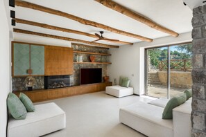 Un salon moderne avec des poutres apparentes en bois, une cheminée en pierre, des étagères intégrées et une grande fenêtre offrant une vue sur un escalier en pierre et la verdure.