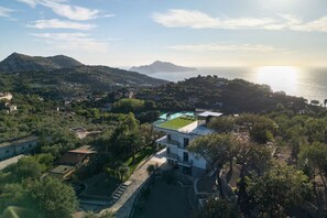 Une vue aérienne d’une ville côtière, où l’on aperçoit un imposant bâtiment blanc doté d’un toit végétalisé, entouré d’une végétation luxuriante et offrant une vue dégagée sur la mer et les montagnes lointaines.
