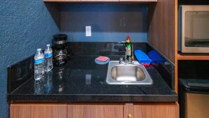 A compact kitchen with a sink, a coffee maker, two water bottles, a dish soap bottle, and a blue sponge.