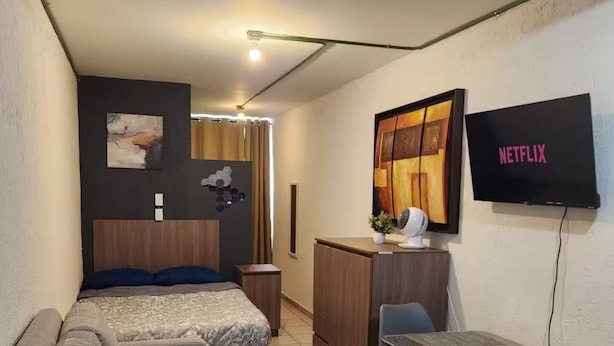 A compact hotel room with a bed, a sofa, a small desk with a chair, a TV, and a wall-mounted artwork.