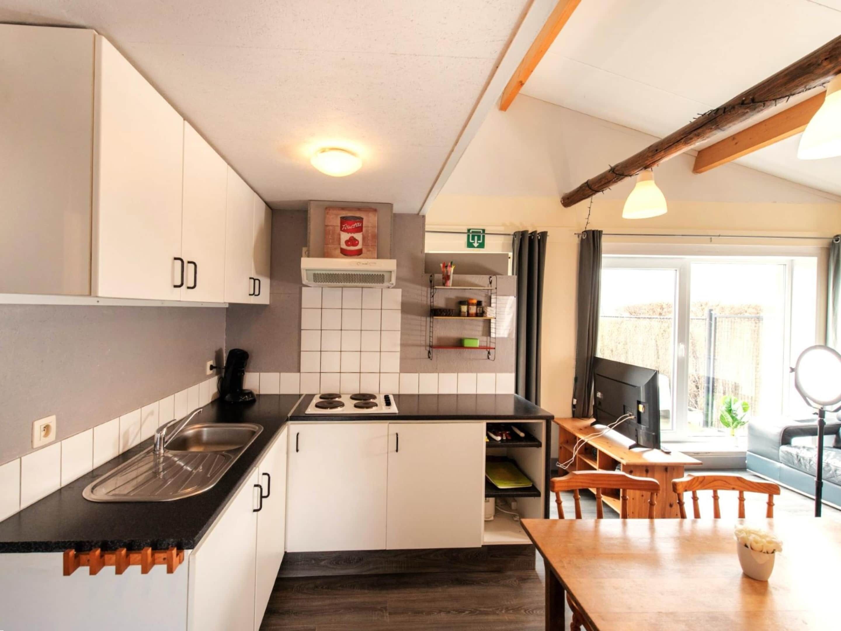 Eine kleine Küche mit weißen Schränken, einer schwarzen Arbeitsplatte und einem eingebauten Herd. Es gibt einen Essbereich mit einem Holztisch und Stühlen, sowie ein Fenster, das natürliches Licht hereinlässt.
