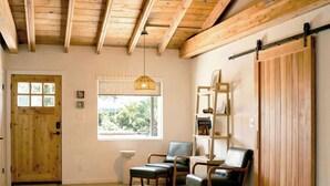Una habitación con vigas de madera, una puerta corrediza estilo granero, un ventanal con vistas, dos sillas, una mesita y una lámpara colgante.