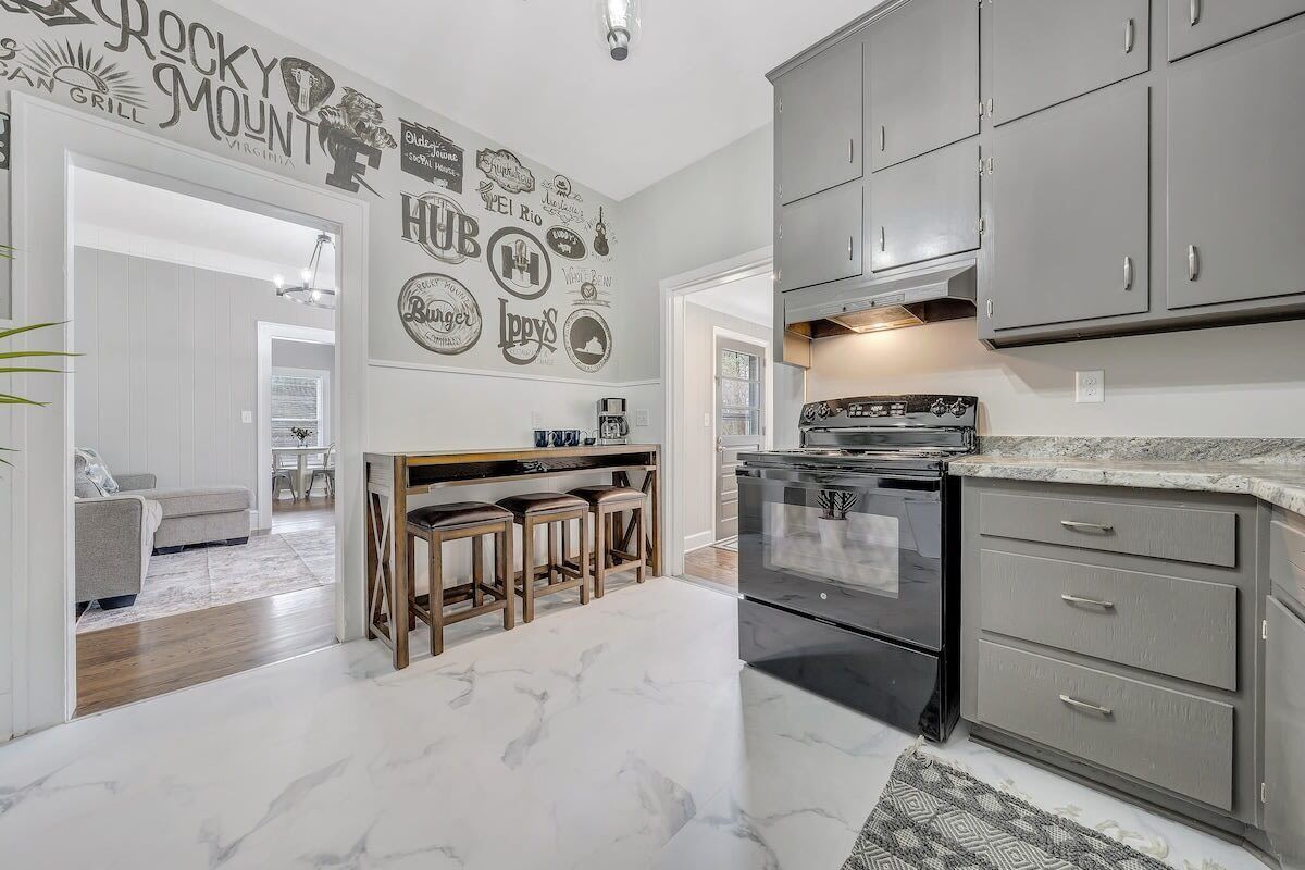 A kitchen with grey cabinets, a black stove, and a marble floor. There is a bar area with stools and a wall decorated with various logos.
