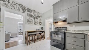 A kitchen with grey cabinets, a black stove, and a marble floor. There is a bar area with stools and a wall decorated with various logos. - Cottage in the Pines (Rocky Mount)