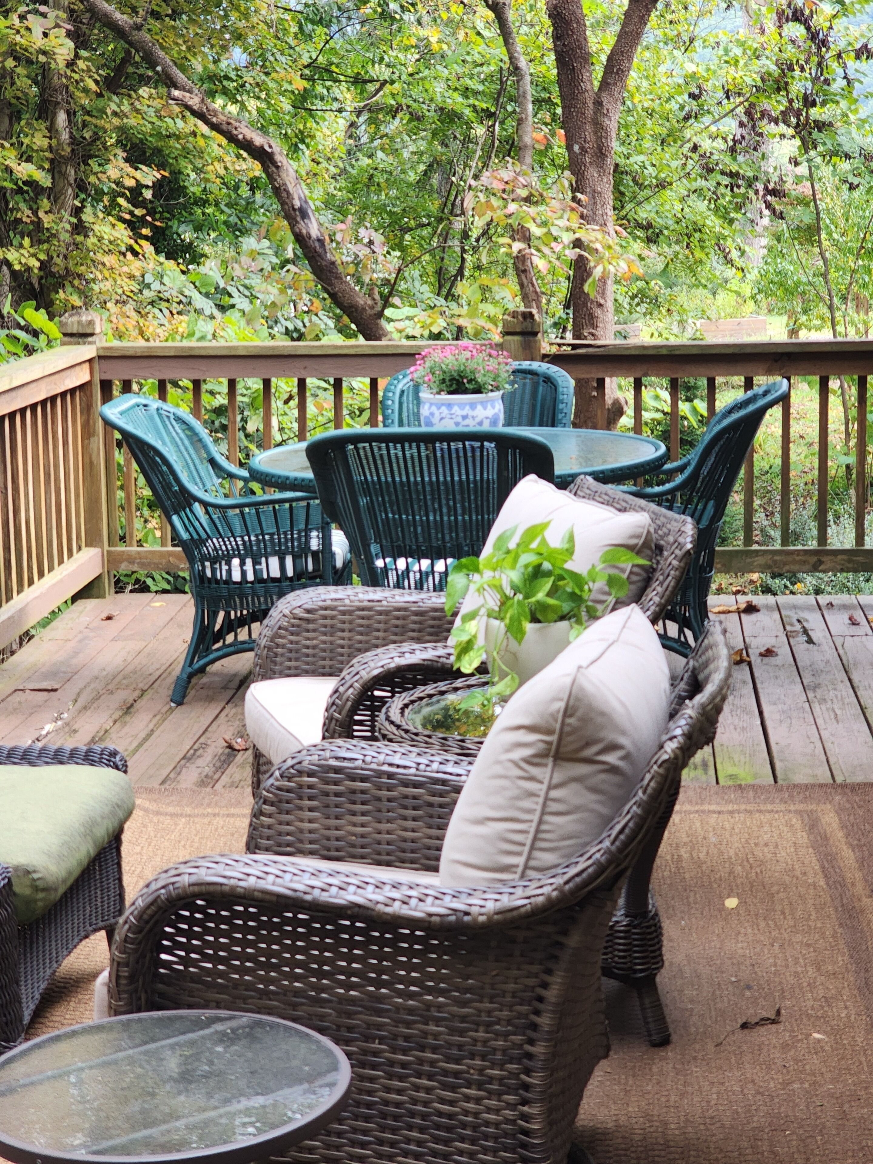 A deck with wicker furniture, including chairs and a table, surrounded by lush greenery.