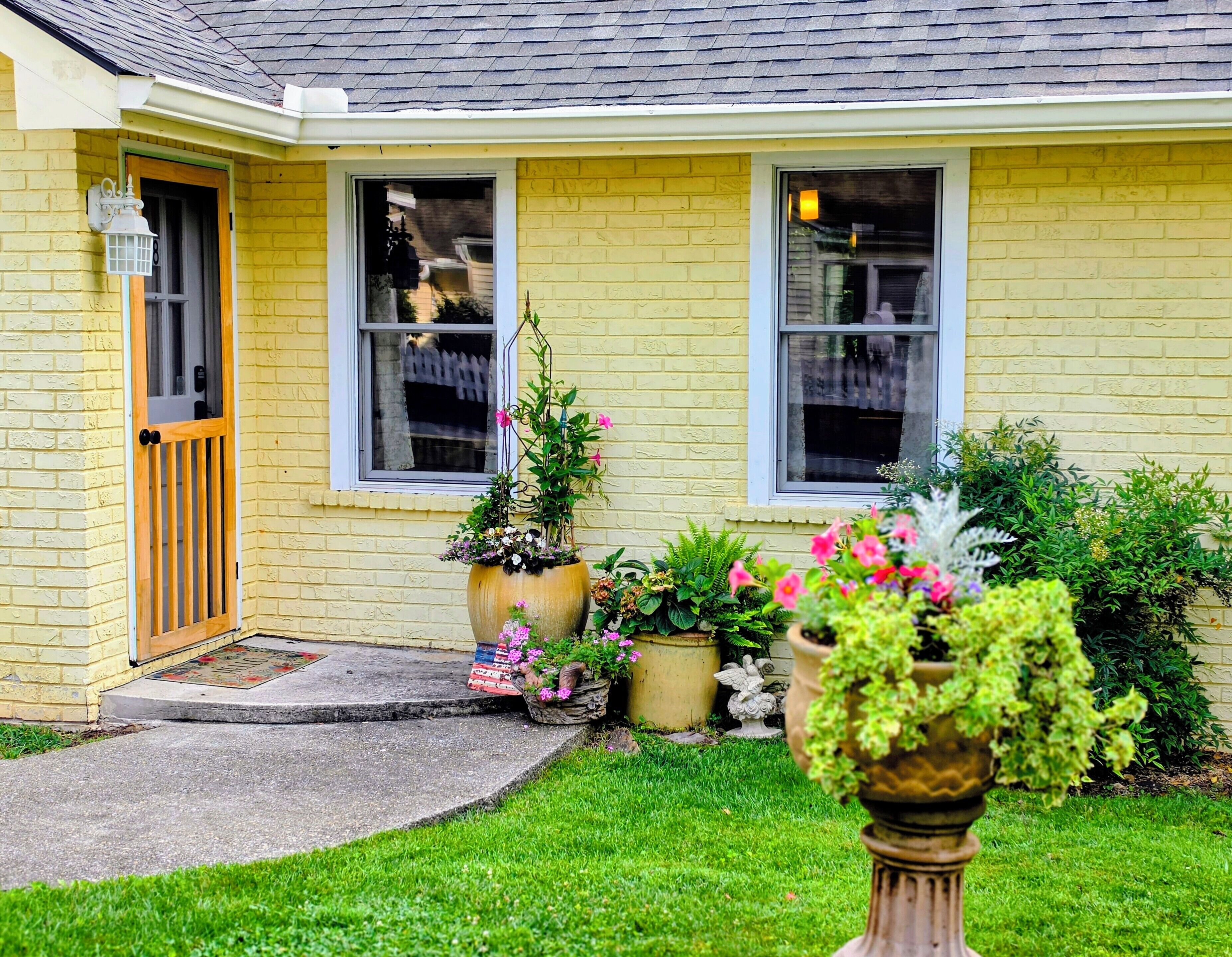 A yellow brick house with a front door, two windows, and a small front yard with potted plants.