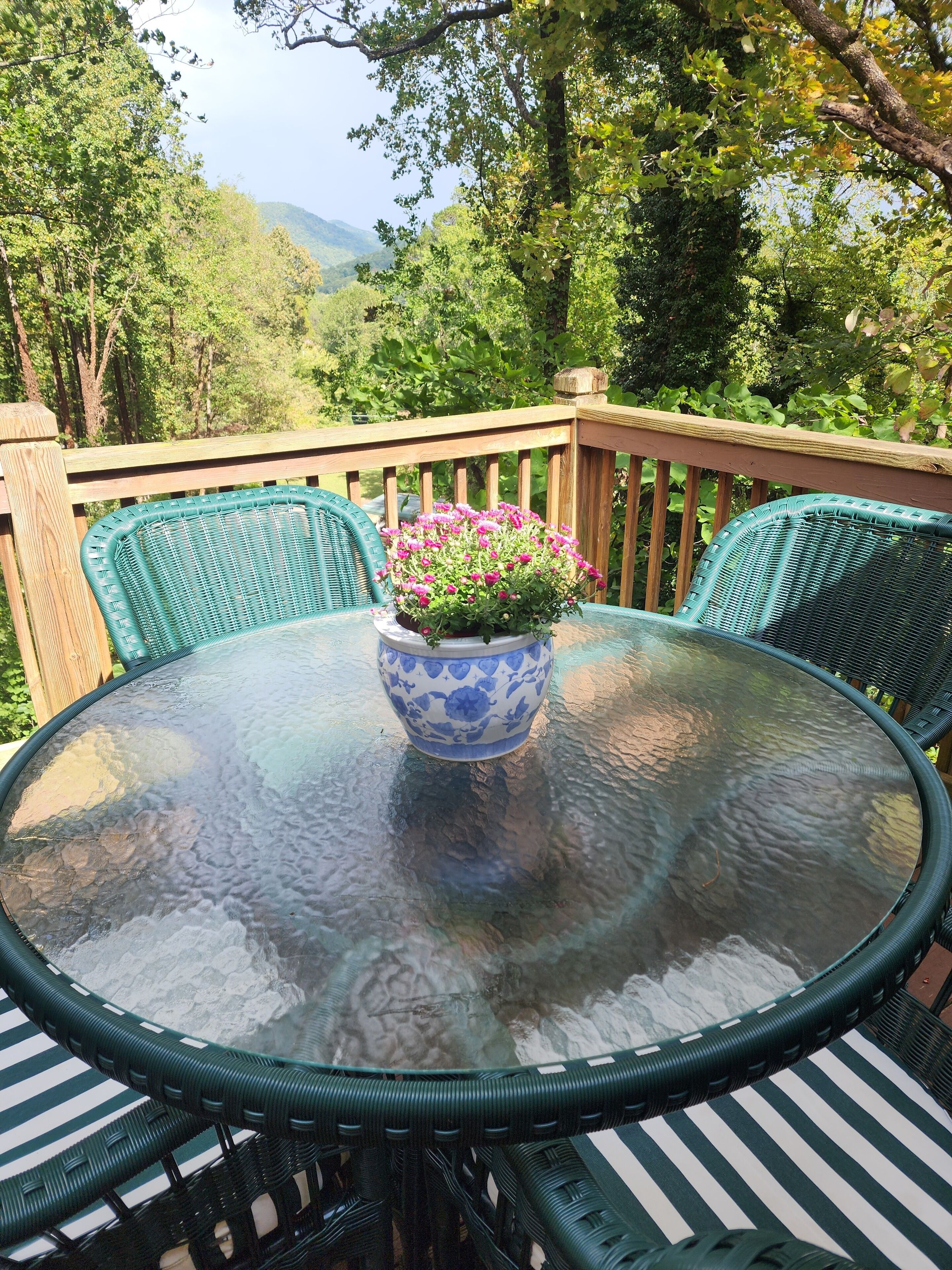 A round glass table with a blue floral vase, surrounded by green wicker chairs on a wooden deck with a view of trees and hills.