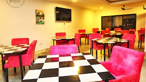 A dining area with red chairs and black-and-white checkered tables, a flat-screen TV on the wall, and ceiling fans. - The Haven Sun N' Fun Hotel (Tanji)