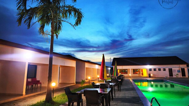 An outdoor pool area with tables and chairs, a palm tree, and a building with illuminated windows. - The Haven Sun N' Fun Hotel (Tanji)