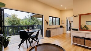 A spacious living area with a dining table, chairs, a kitchen, and a balcony with a view of greenery. - Hidden Springs Retreat - Karangahake Mountain View (Waihi)