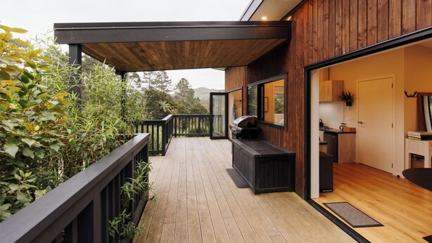 A wooden deck with a built-in barbecue area, a kitchen visible through an open door, and a view of greenery and hills in the background. - Hidden Springs Retreat - Karangahake Mountain View (Waihi)