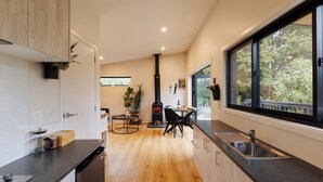 A modern kitchen with a central island, a built-in stove, and a large window providing a view of the outdoors. - Hidden Springs Retreat - Karangahake Mountain View (Waihi)