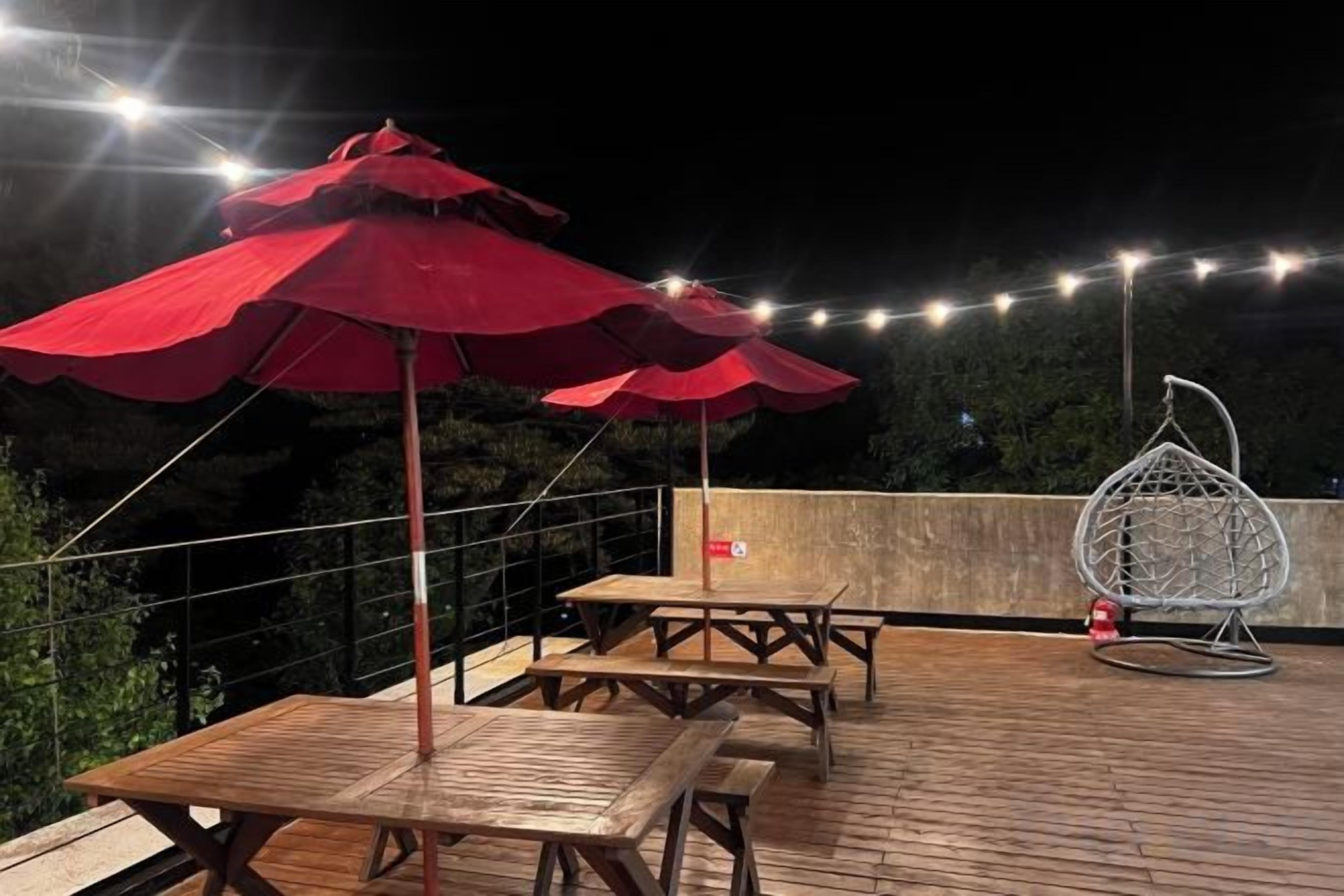 Een terras buiten met houten tafels en rode parasols, een hangstoel en lichtjes.