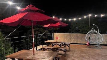 Een terras buiten met houten tafels en rode parasols, een hangstoel en lichtjes.