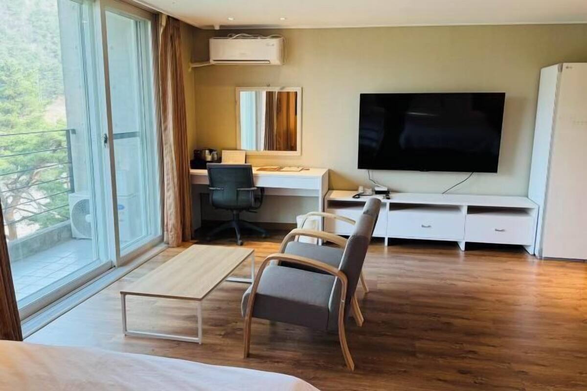 Quarto de hotel com uma janela grande, uma mesa, uma cadeira, uma televisão e um guarda-roupa.