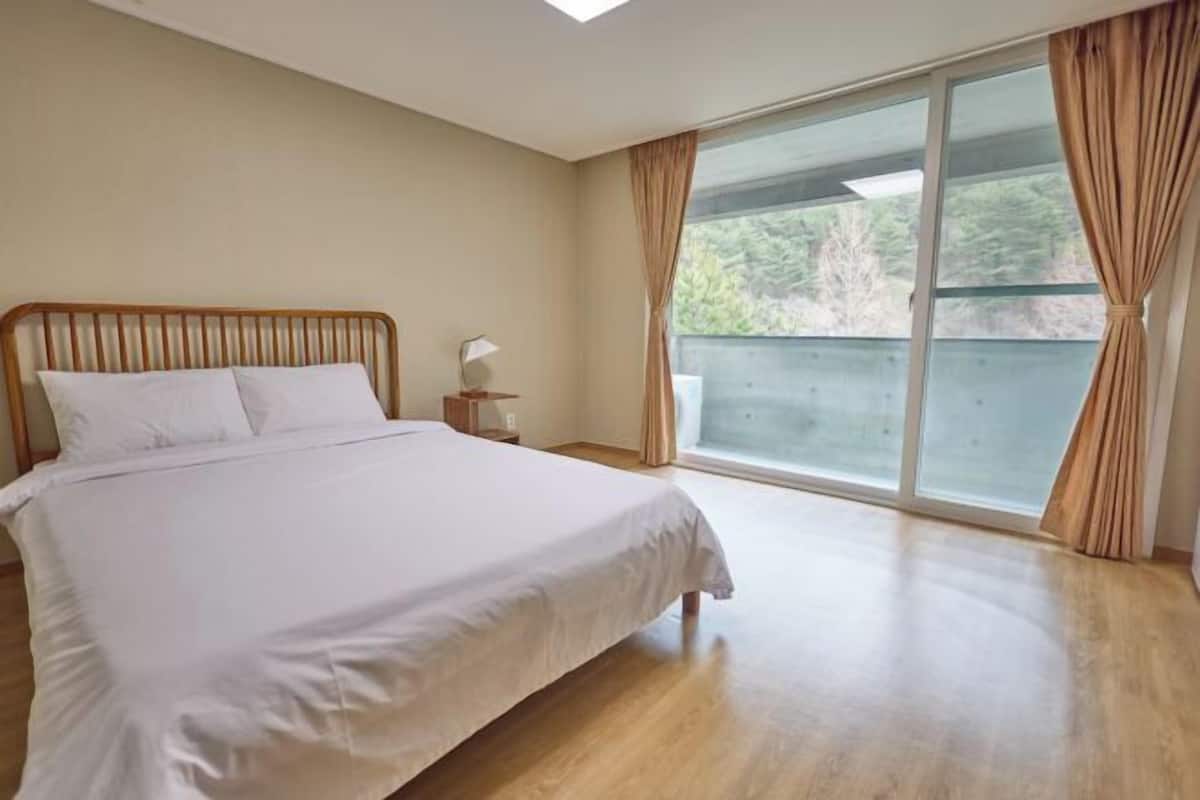 Quarto com cama, mesa de cabeceira e uma janela grande com cortinas.