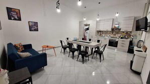 Un saló modern amb un sofà blau, una taula de menjador amb cadires i una cuina amb armaris blancs i una façana de rajola.