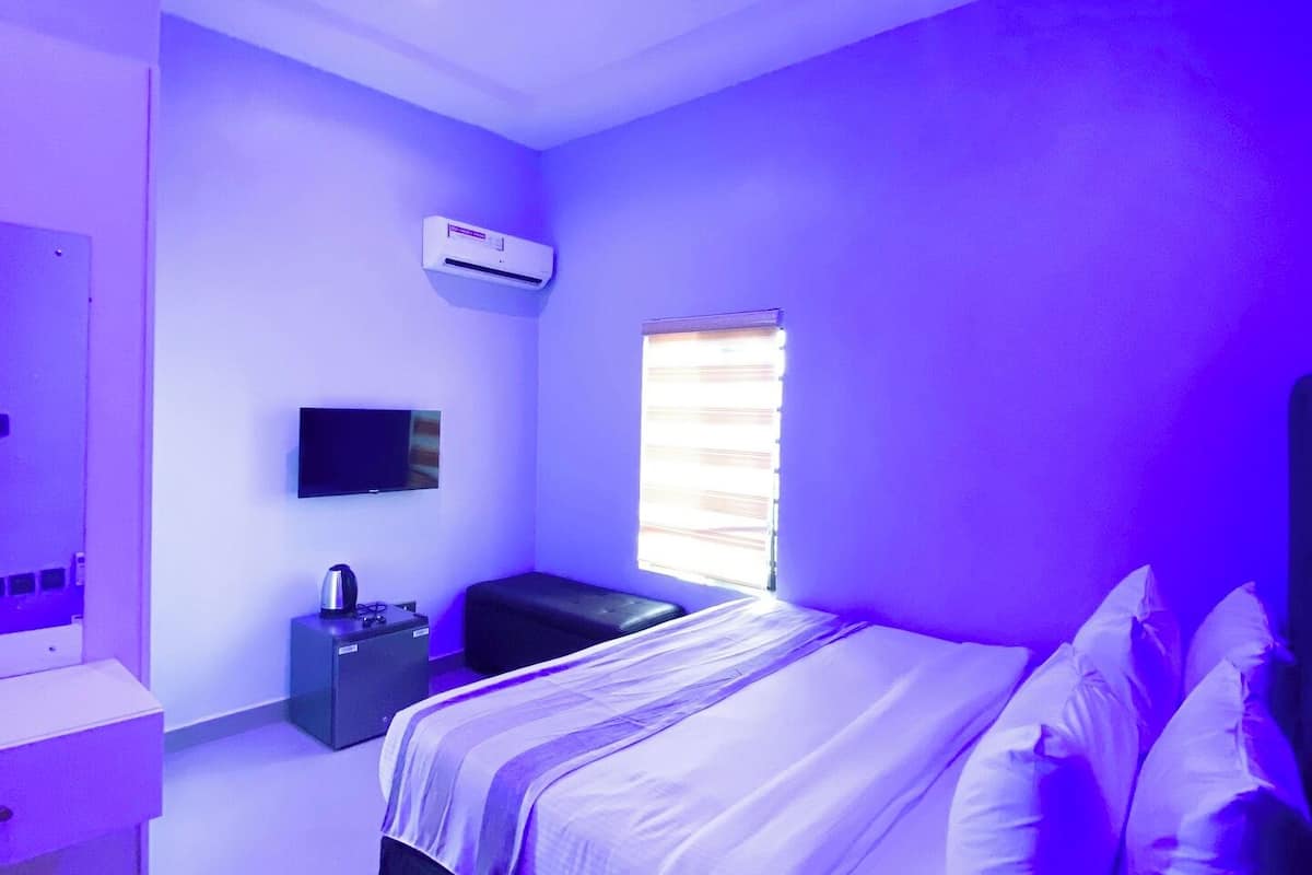 Une chambre moderne avec un lit, une télévision, un petit bureau et une fenêtre avec des stores.