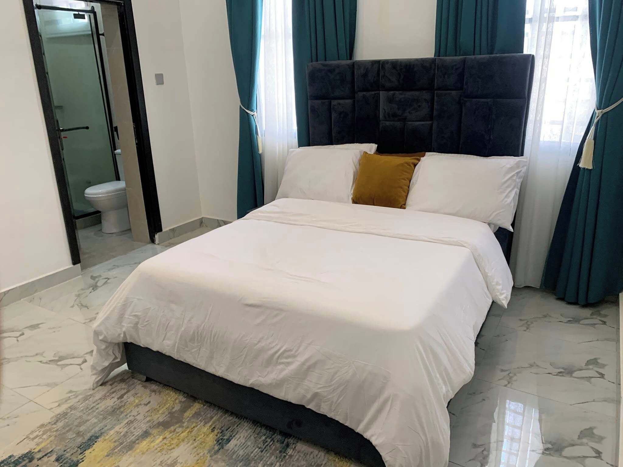 A neatly made bed with white bedding and a dark headboard, a bathroom with a toilet visible, and teal curtains.