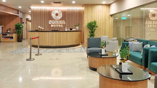 A hotel reception area with a wooden reception desk, a seating area with a sofa and armchairs, and a glass wall. - Cordia Hotel Yogyakarta - Airport Hotel (Temon)