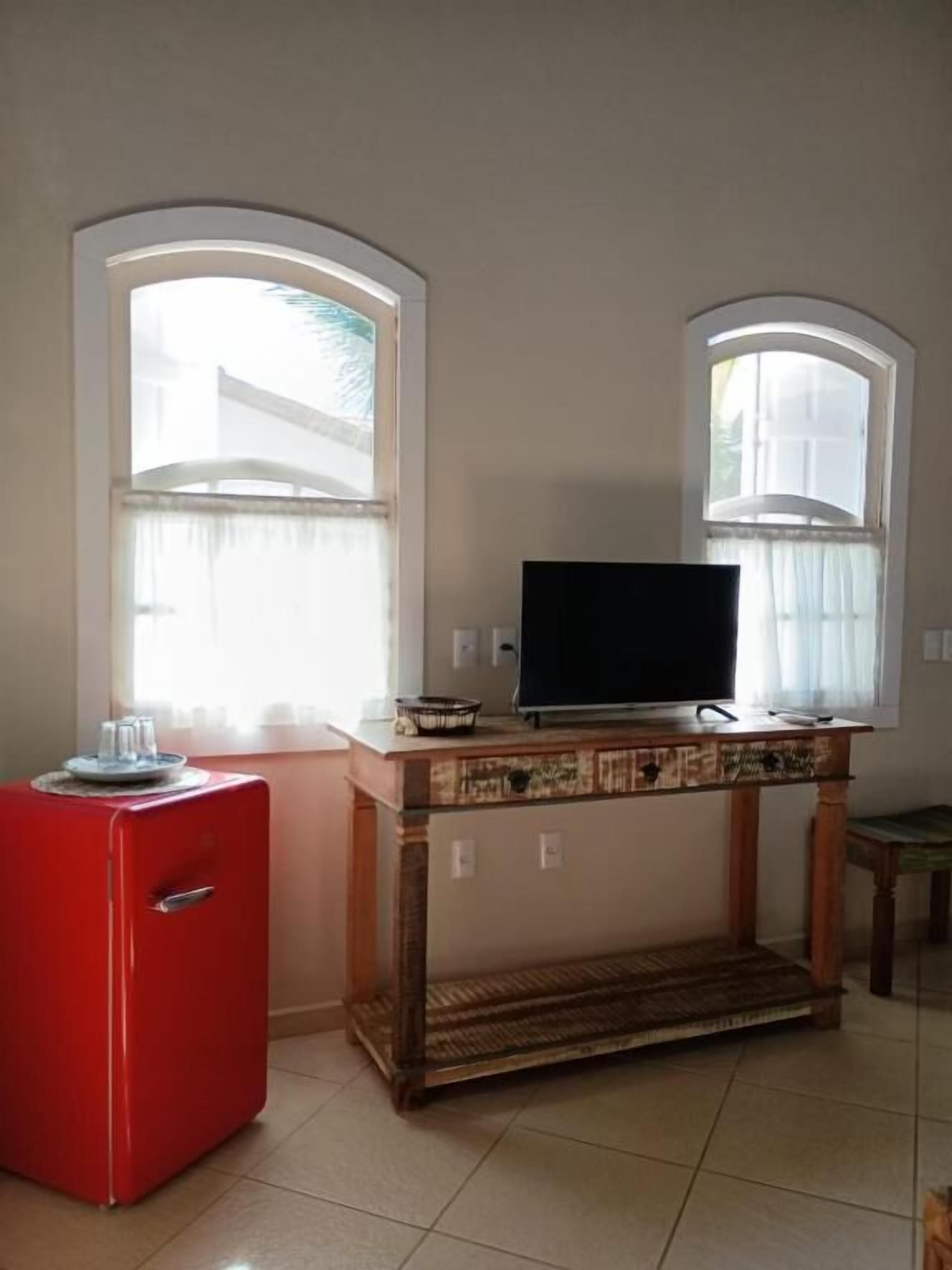 A small room with a television on a wooden stand, a red appliance, and two windows with curtains.