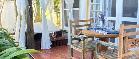 Un porche cubierto con una mesa y sillas de madera, una ventana con cortinas y una zona con plantas.
