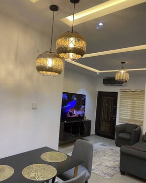 A living room with a TV, two hanging lamps, a sofa, and a dining table with placemats. - Konar Apartment (Abuja)