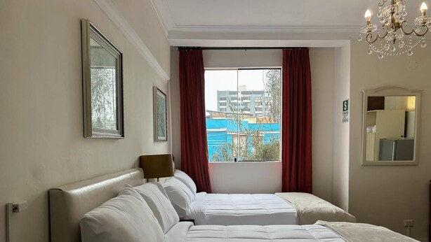 Ein Hotelzimmer mit zwei Betten, einem großen Fenster mit roten Vorhängen, einer Kristallleuchtre und Blick auf Gebäude.